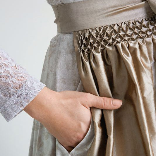 Detailaufnahme einer Hand, die in die Tasche eines elegant gefalteten Kleides greift, mit einem Spitzenaufschlag am Ärmel.