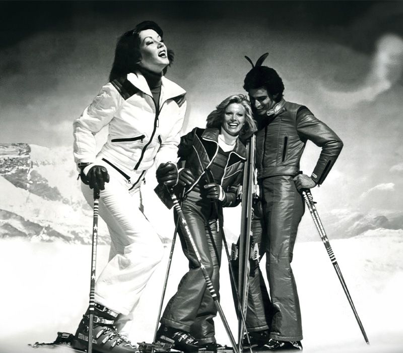 Drei Skifahrer in stylischer Ski-Bekleidung posieren fröhlich auf einer verschneiten Berglandschaft.