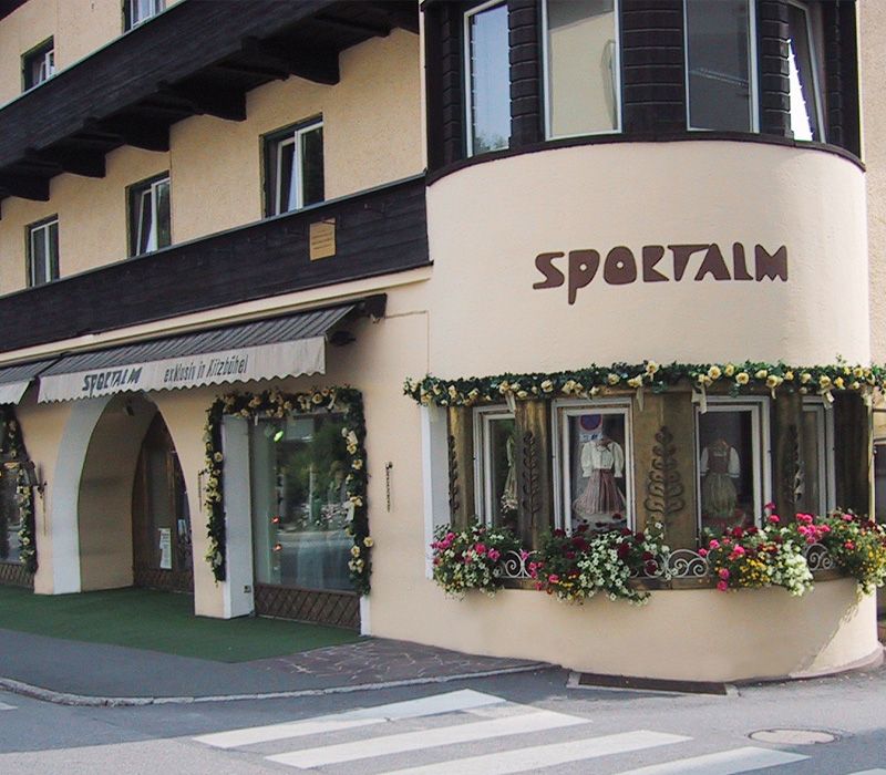 Fassade eines Hauses mit dem Schriftzug "SPORTALM" und blühenden Fensterdekorationen.