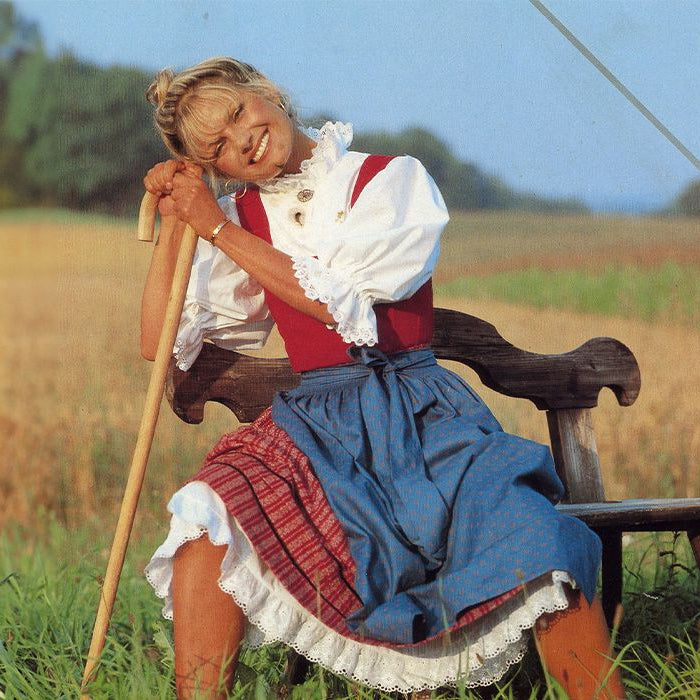 Eine lächelnde Frau in traditioneller Tracht sitzt auf einer Bank in einem Feld.