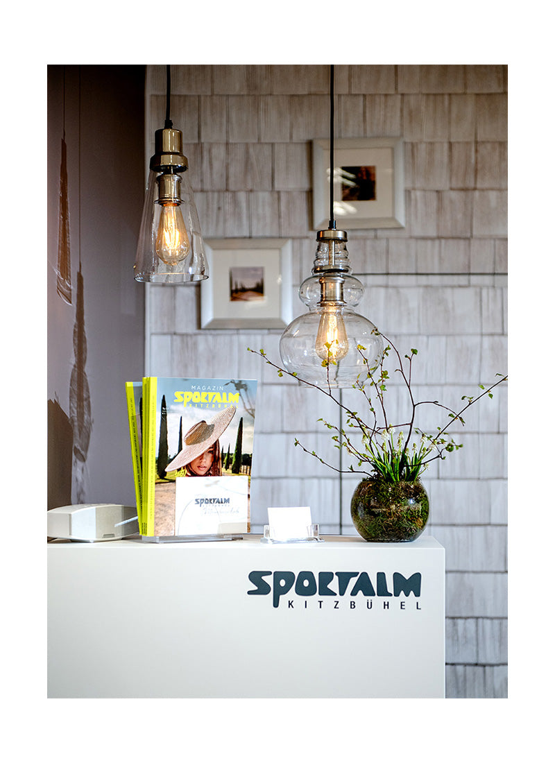 Eine moderne Empfangstheke mit einem Buch über Sportalm, einer Vase mit Pflanzen und zwei hängenden Lampen.