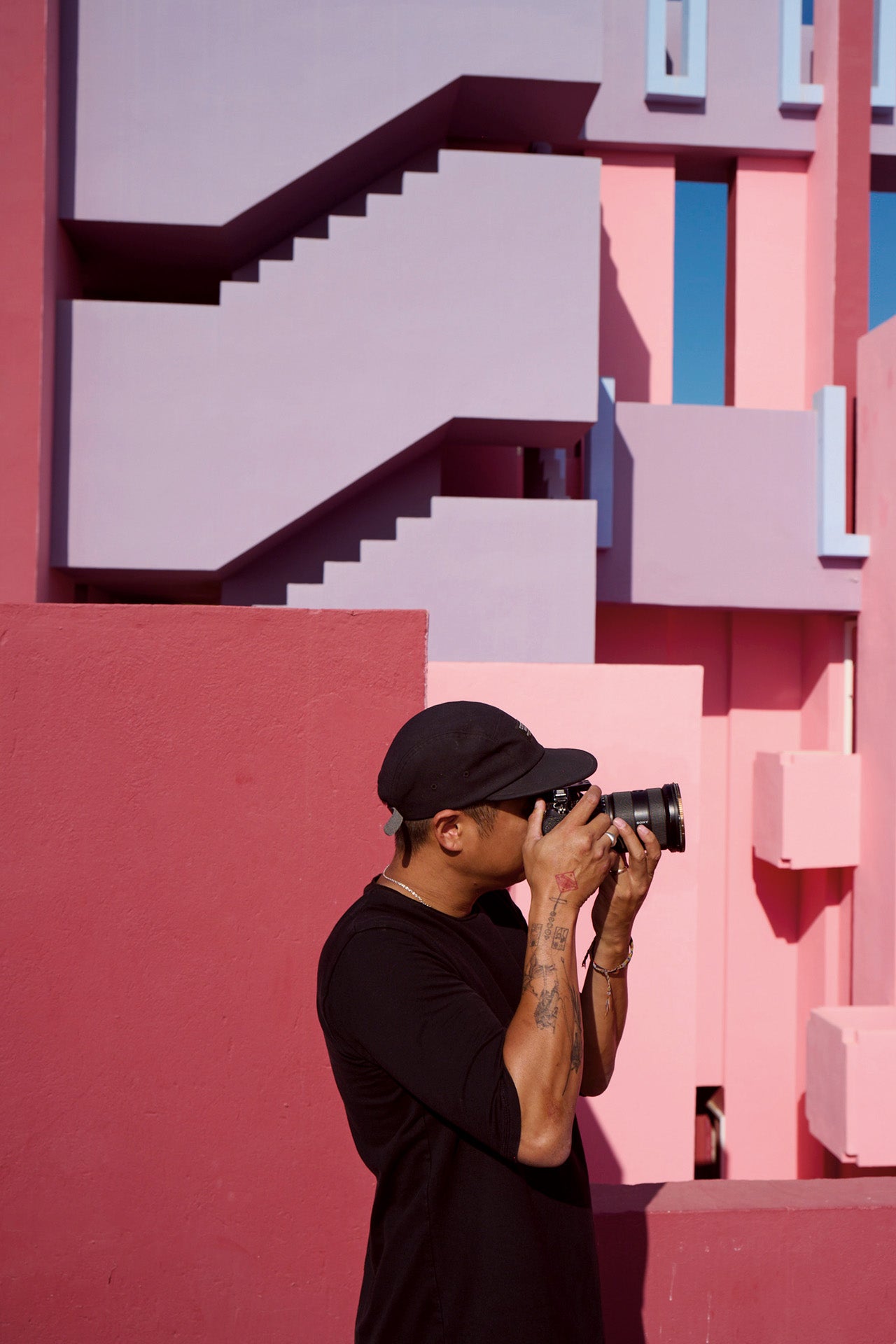 Ein Mann mit einer Kamera steht vor einer mauerfarbenen Architektur mit treppenförmigen Elementen in Pink- und Lila-Tönen.