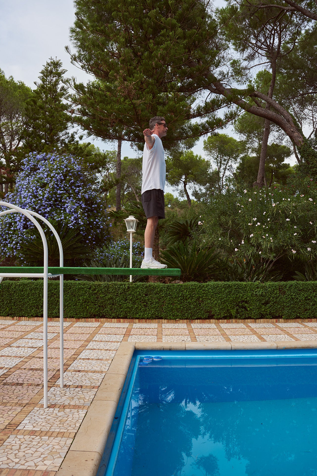 Ein Mann steht auf einem Sprungbrett neben einem Pool, umgeben von Bäumen und Blumen.