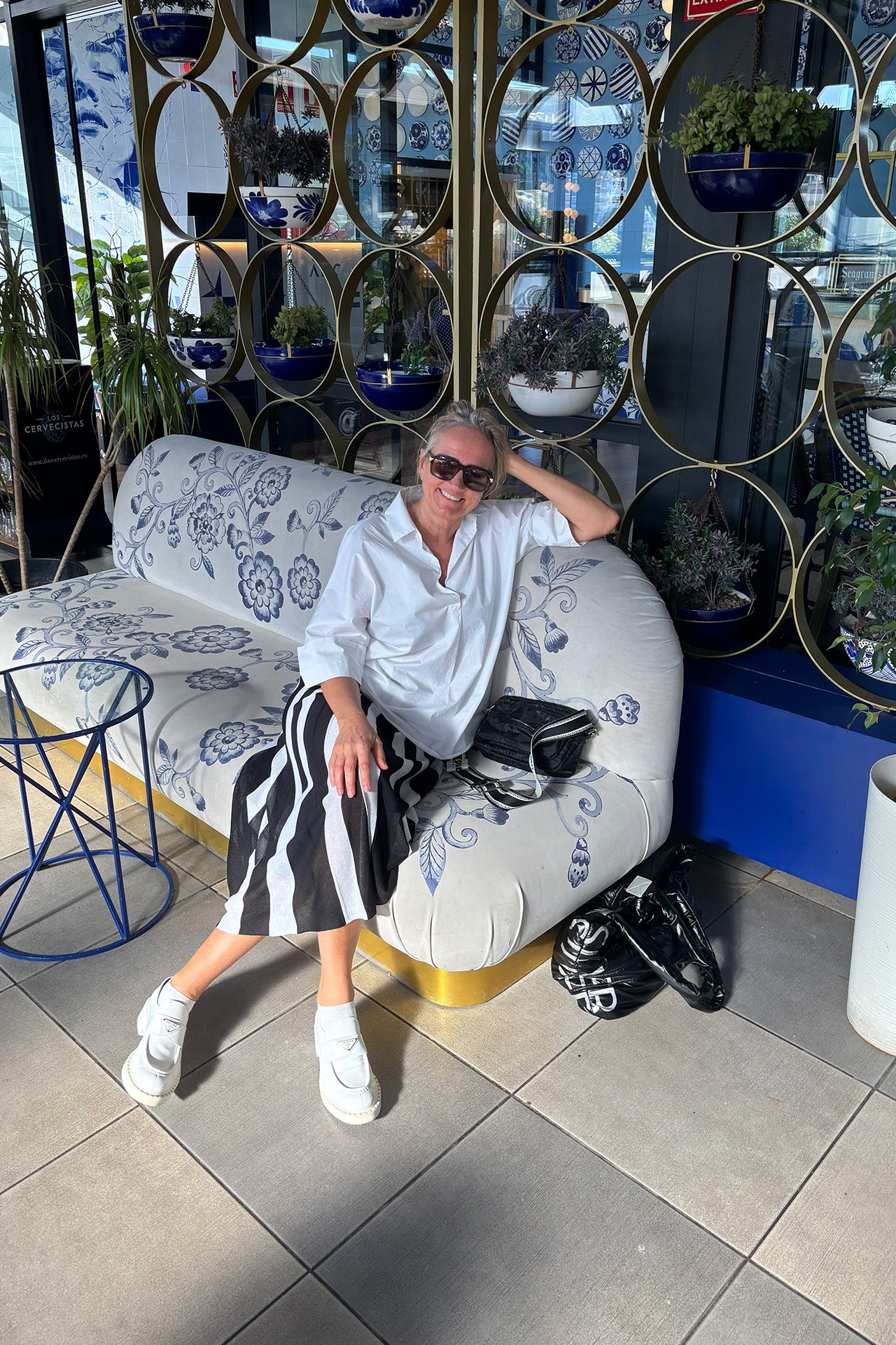 Frau mit Sonnenbrille sitzt entspannt auf einem blauen Sofa, trägt ein weißes Hemd und einen schwarz-weißen Rock, umgeben von Pflanzen.