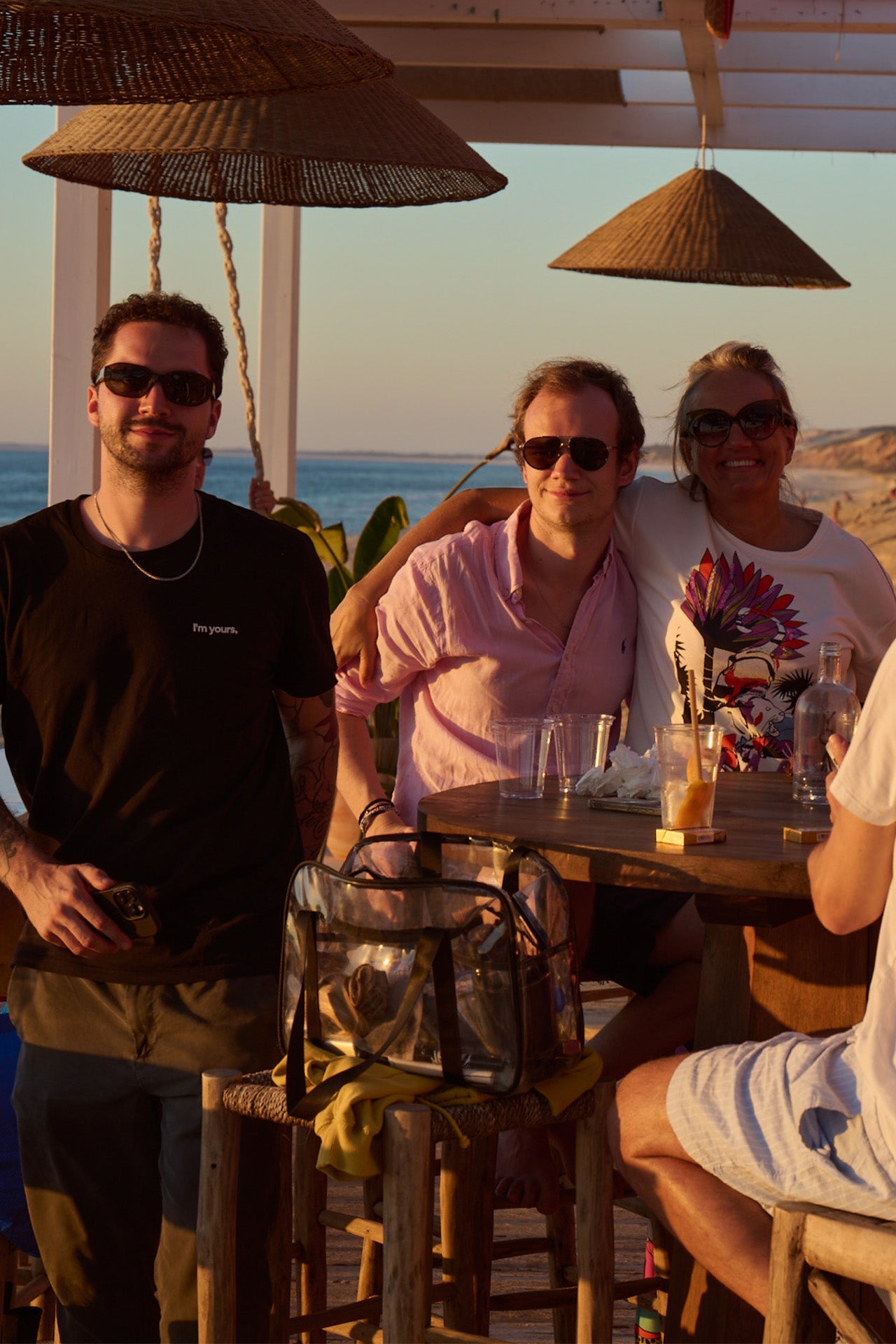 Drei Personen stehen an einem Tisch in einer Strandbar bei Sonnenuntergang, während eine Person an einem Getränk arbeitet.