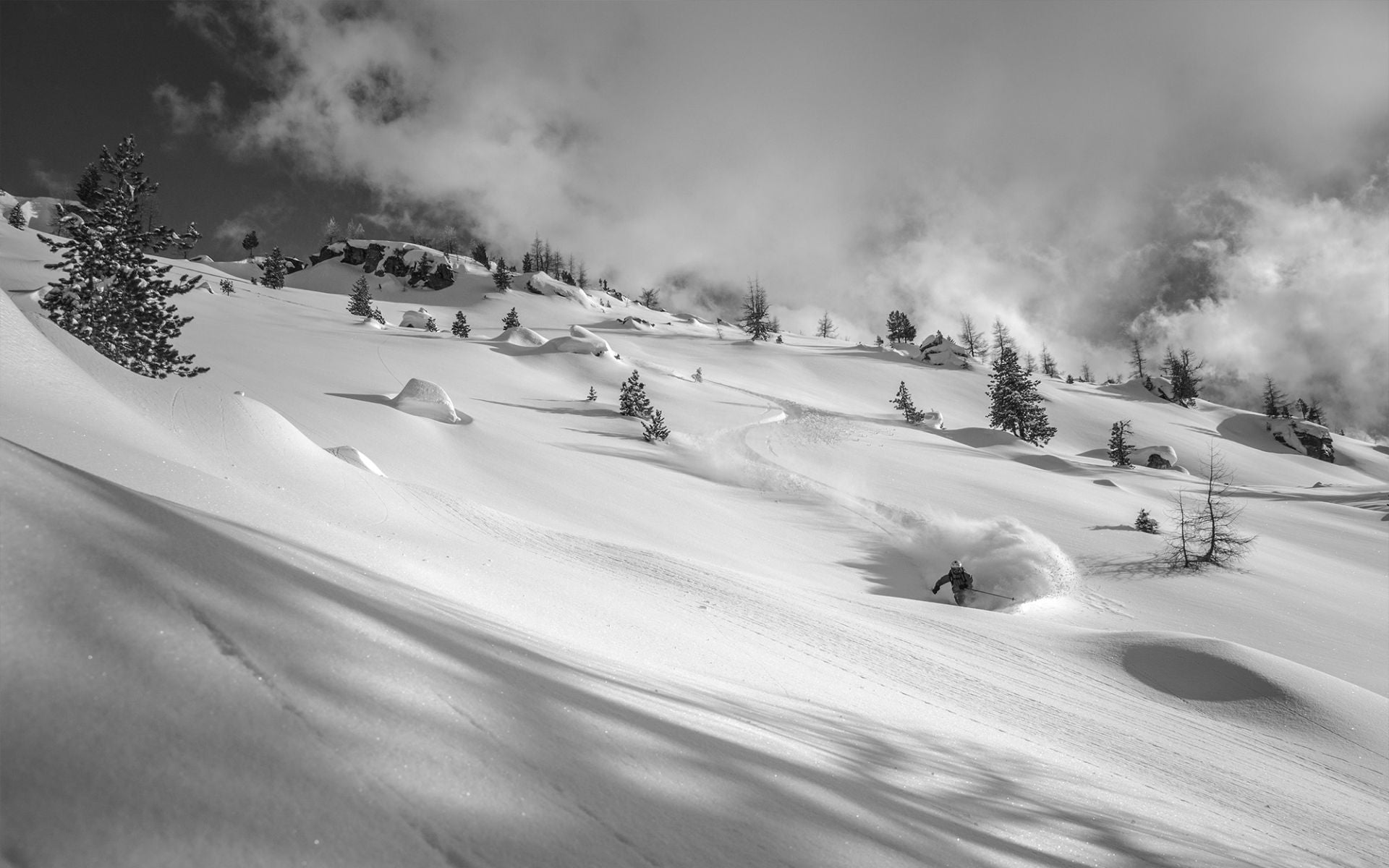 Ein Skifahrer fährt durch unberührten, tiefen Schnee in einer winterlichen Landschaft mit bewaldeten Hügeln und wolkigem Himmel.