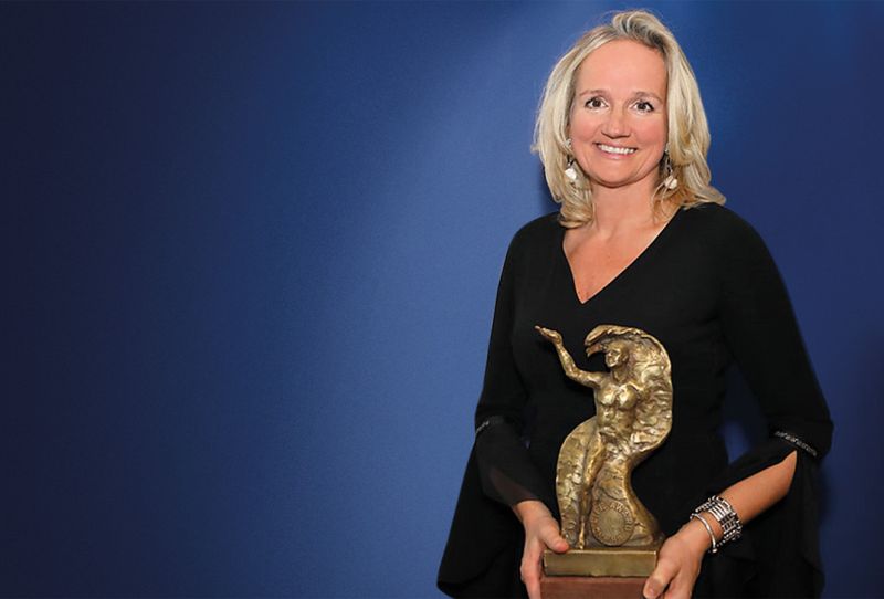 Frau mit blonden Haaren hält eine goldene Statue in der Hand und lächelt vor einem blauen Hintergrund.