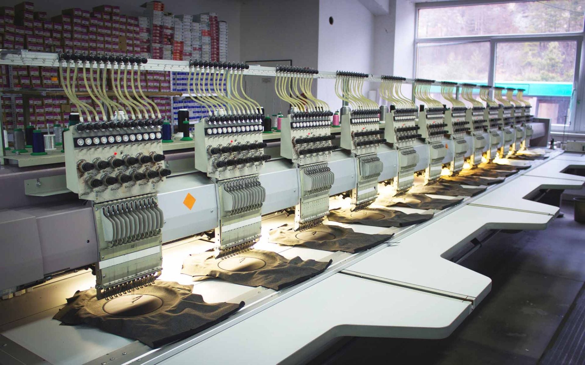 Mehrere industrielle Stickmaschinen stehen in einer Reihe auf einem Tisch, mit beleuchteten Arbeitsflächen und gestickten Stoffen darunter.