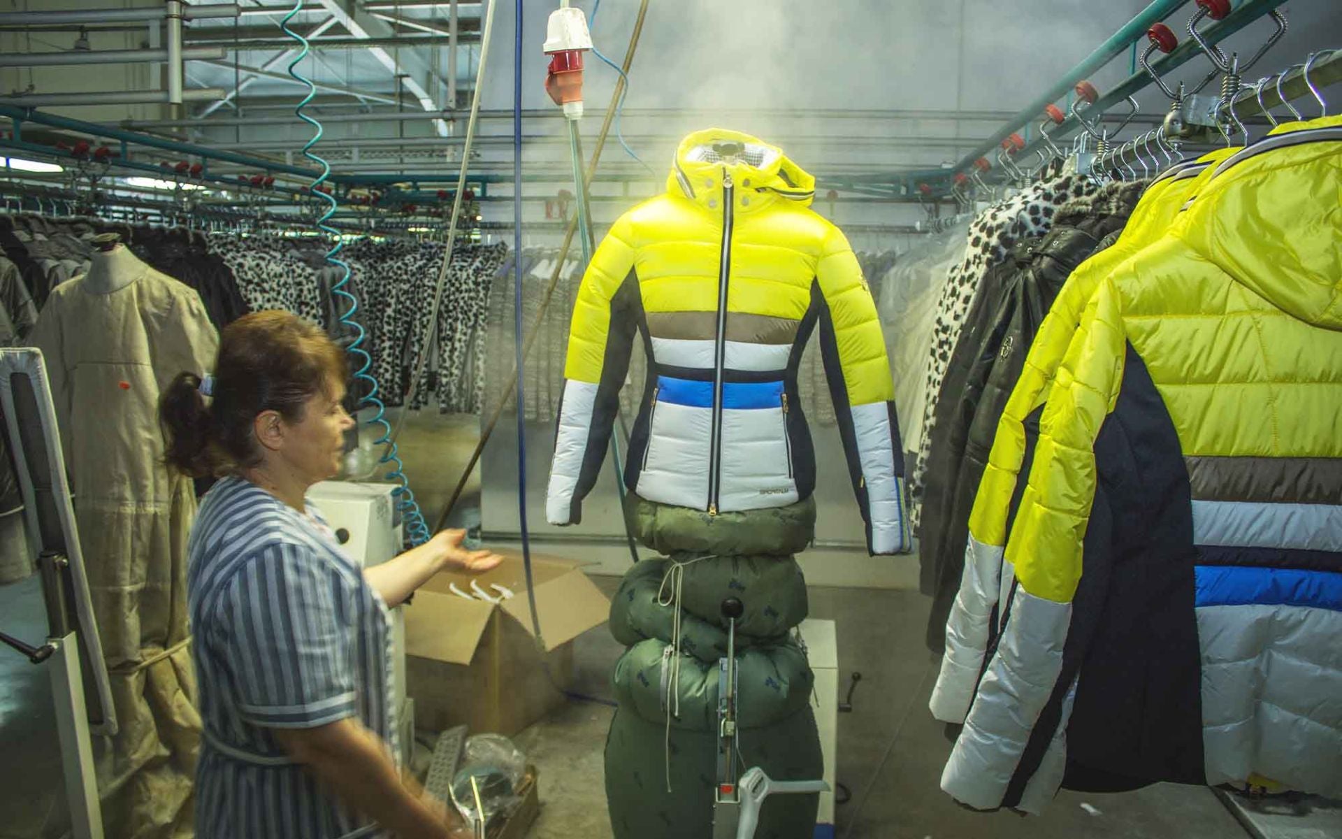 Eine Frau in einer Textilfabrik präsentiert eine leuchtend gelbe, gepolsterte Jacke auf einem Kleiderständer, während Dampf im Raum sichtbar ist.