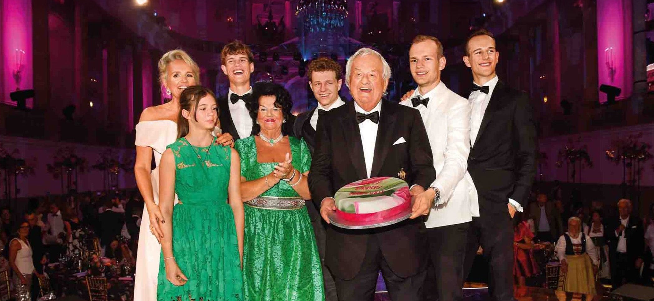 Eine Gruppe von acht Personen in festlicher Kleidung posiert lächelnd um eine große Torte.