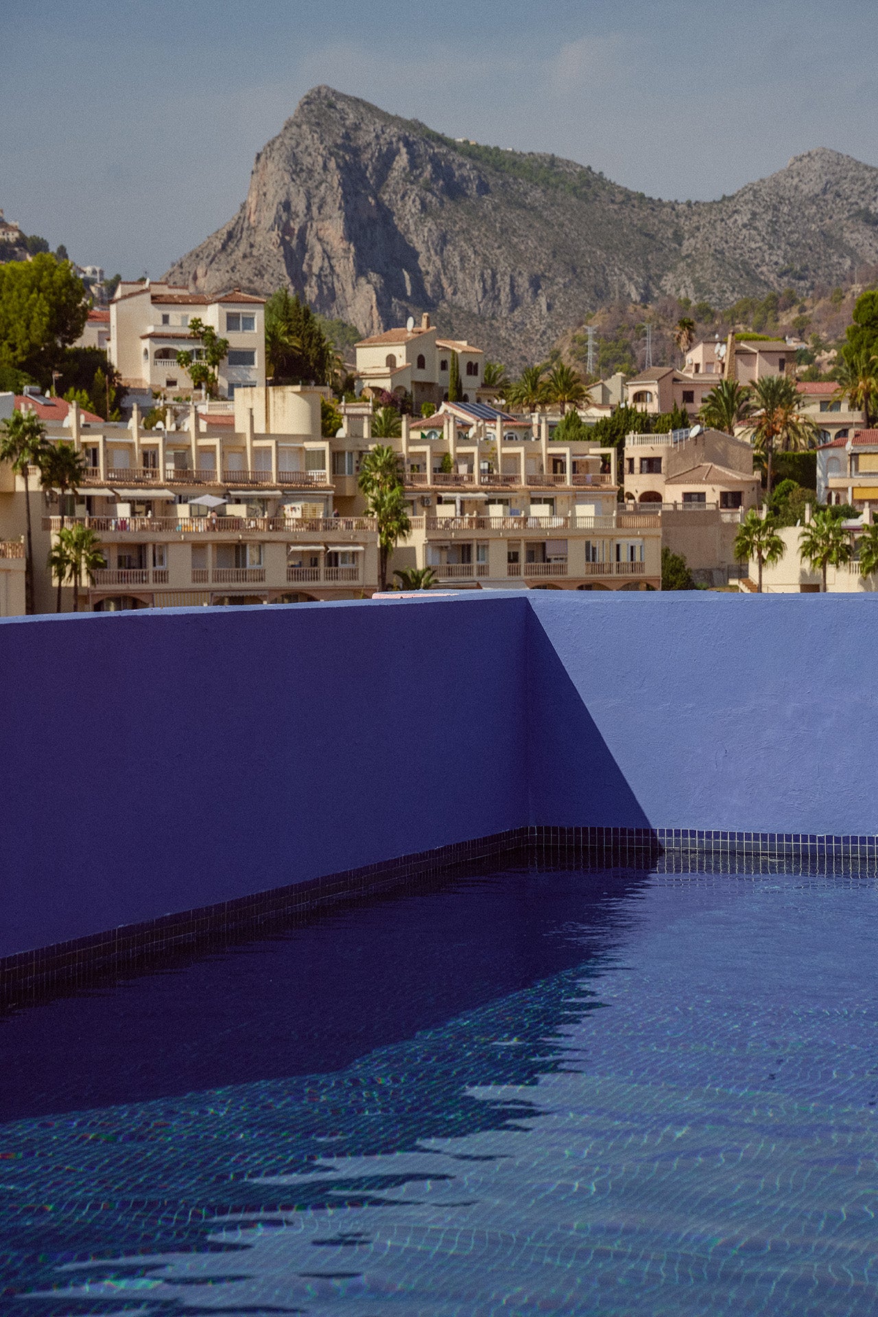 Blick auf eine Poollandschaft mit blauem Wasser, umgeben von einem bunten Wohngebiet und Bergen im Hintergrund.