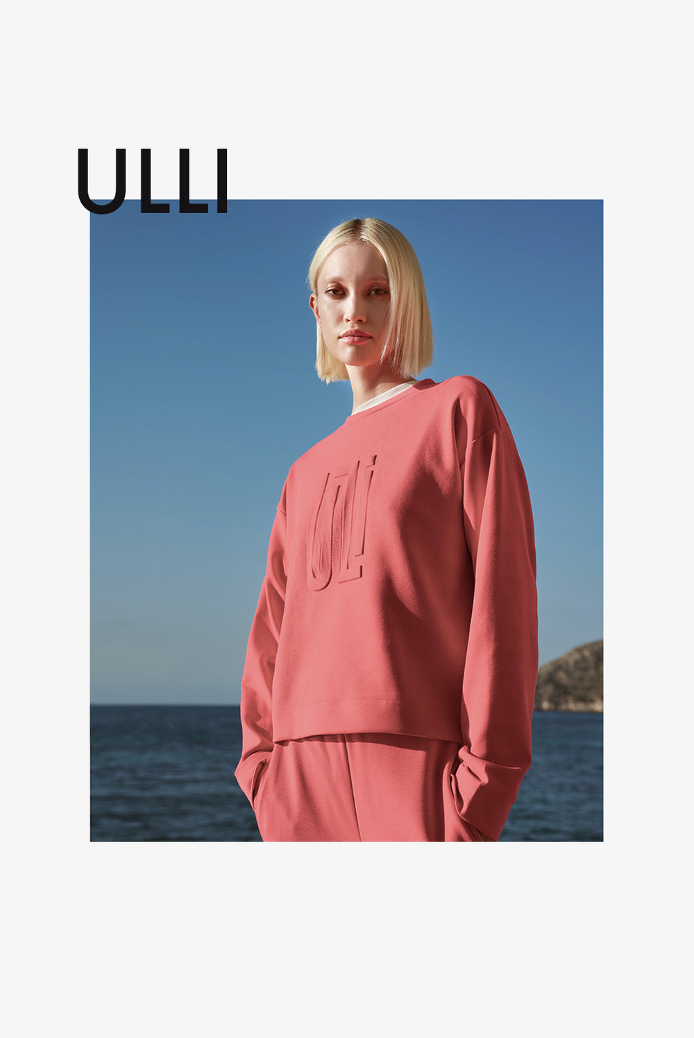 Porträt einer blonden Frau in einem rosanen Pullover mit eingesticktem Schriftzug, vor einem klaren blauen Himmel und Wasser im Hintergrund.