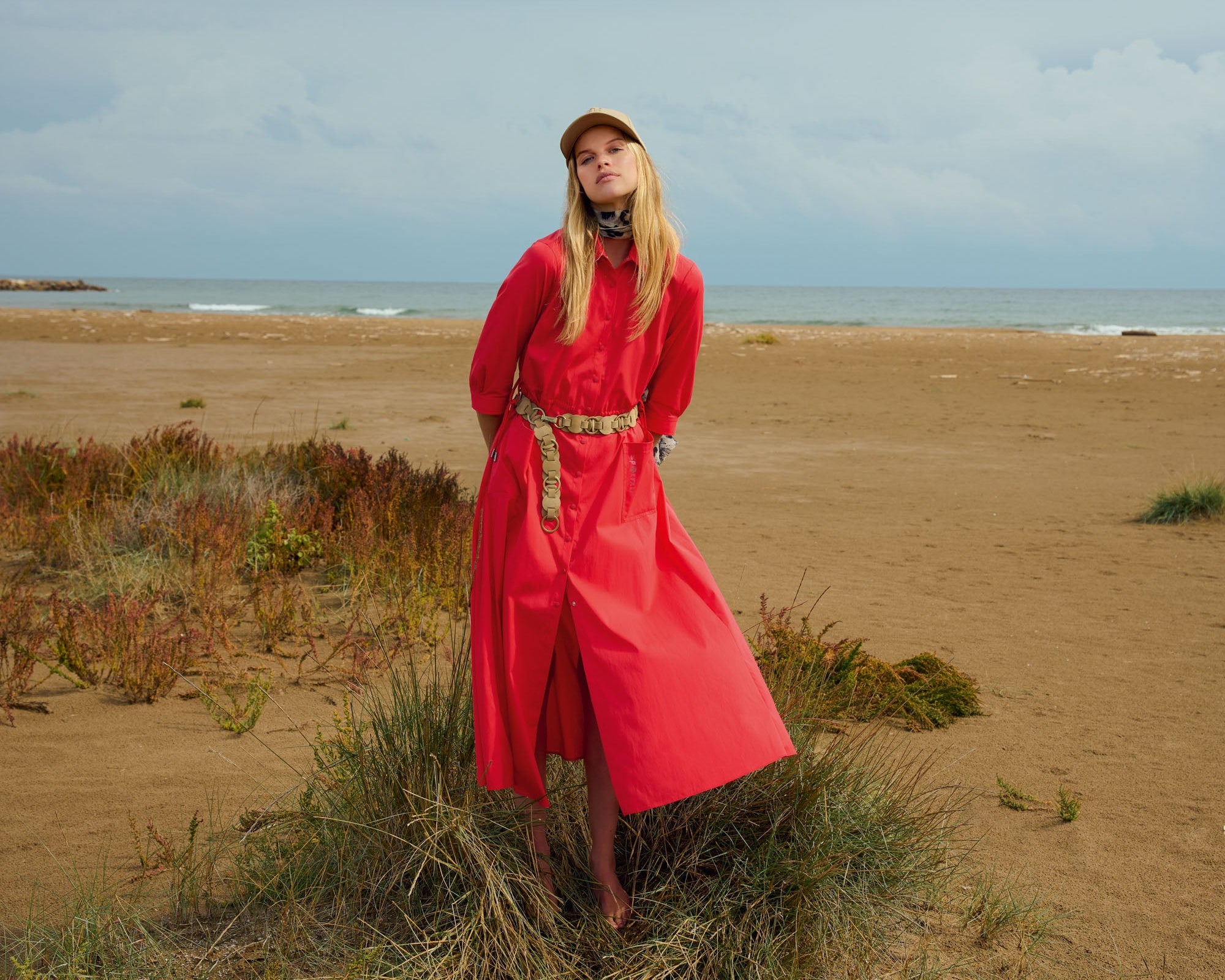 Frau in rotem Kleid und Kappe steht auf einem Sandstrand mit weitem Meer im Hintergrund.