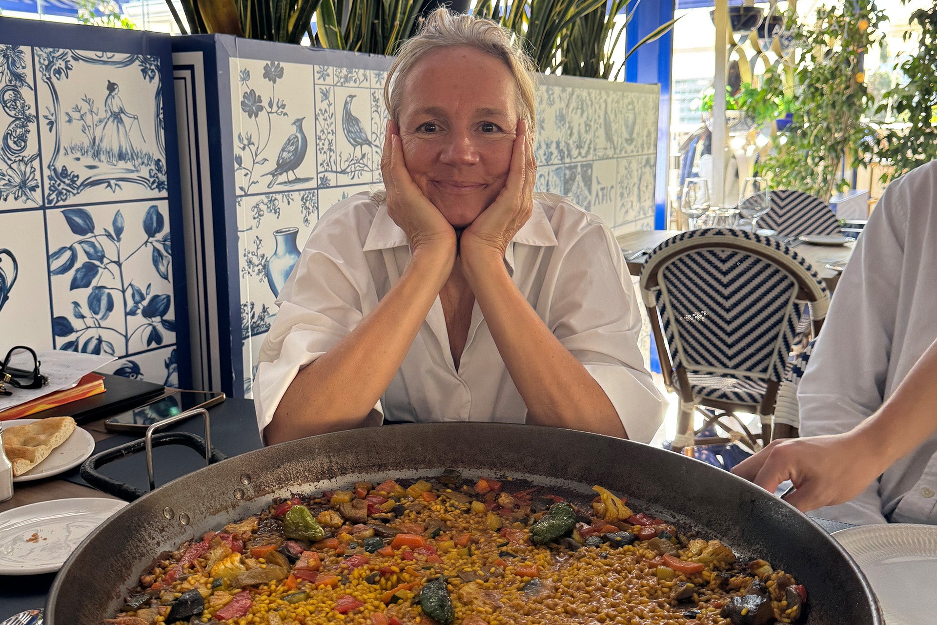 Eine Frau mit kurzen, grauen Haaren sitzt mit einem Lächeln vor einer großen Pfanne Paella in einem hellen Restaurant.