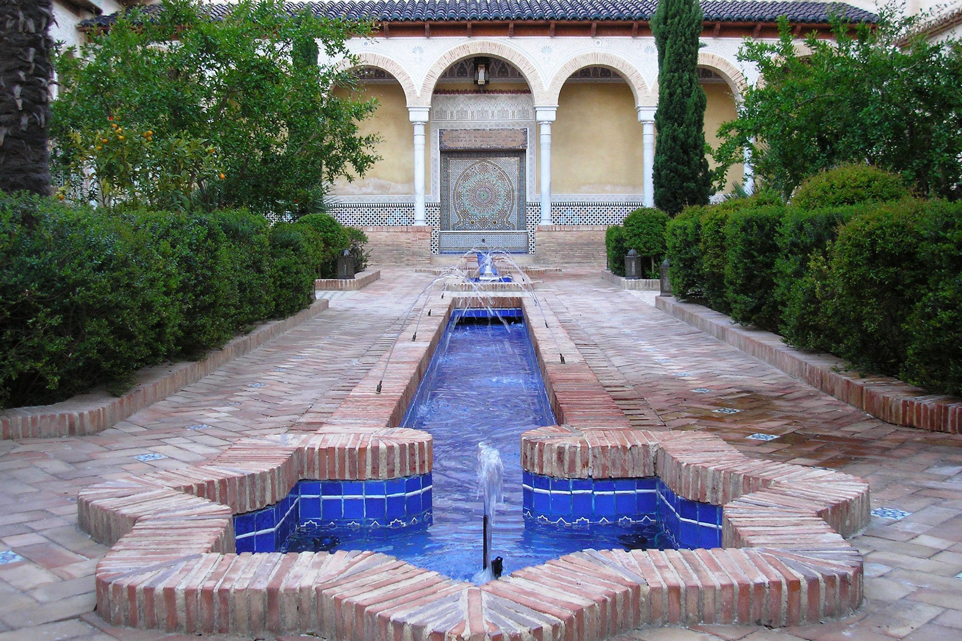 Ein geometrisch angelegter Garten mit einem Wasserlauf und blau gefliesten Elementen, umgeben von strauchförmigen Hecken und einem historischen Gebäudeteil im Hintergrund.
