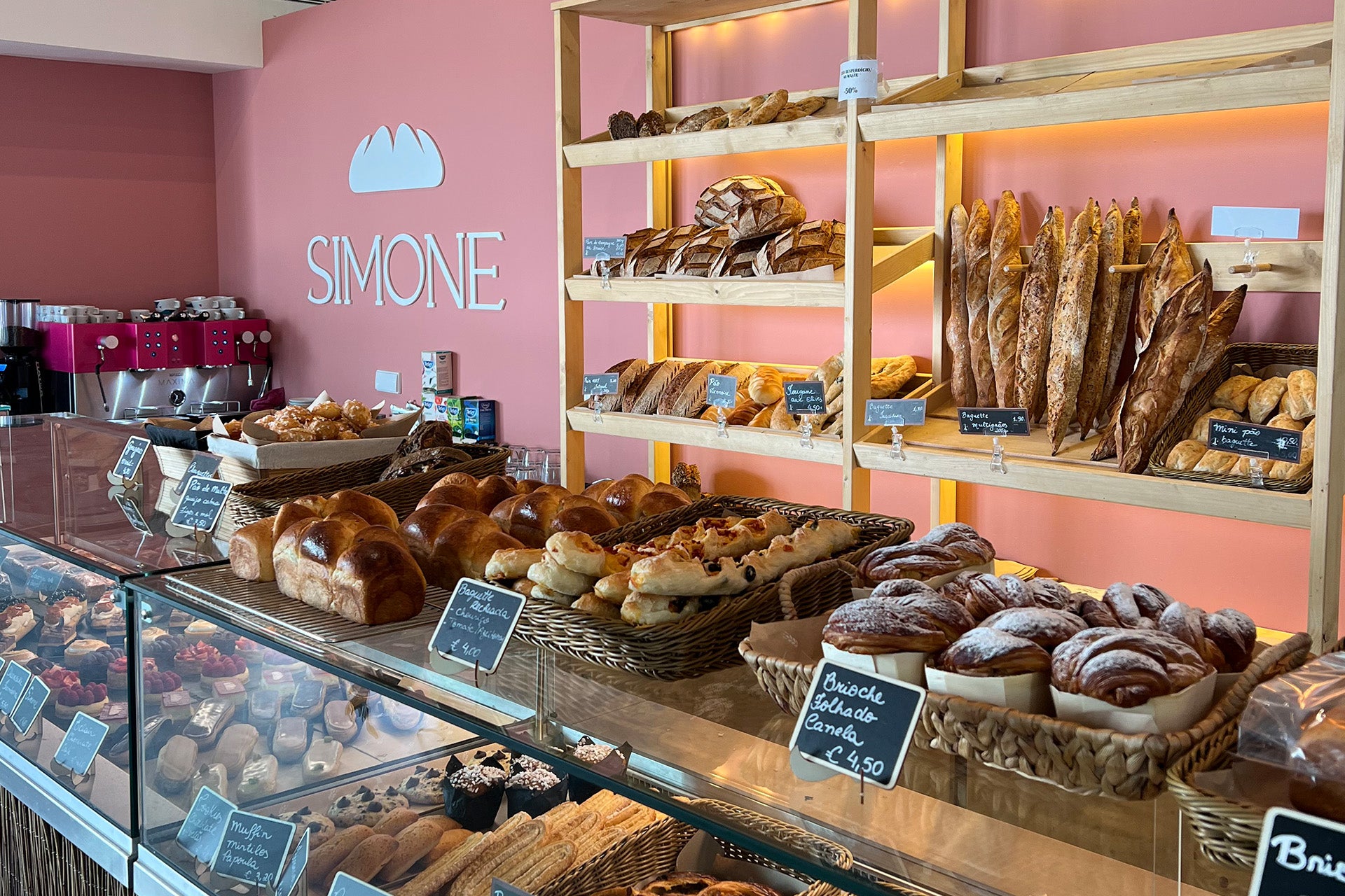 Ein gut gefülltes Cafe mit einer Vitrine voller Backwaren, darunter Brötchen, Croissants und Gebäck, vor einer rosa Wand mit dem Schriftzug "SIMONE".