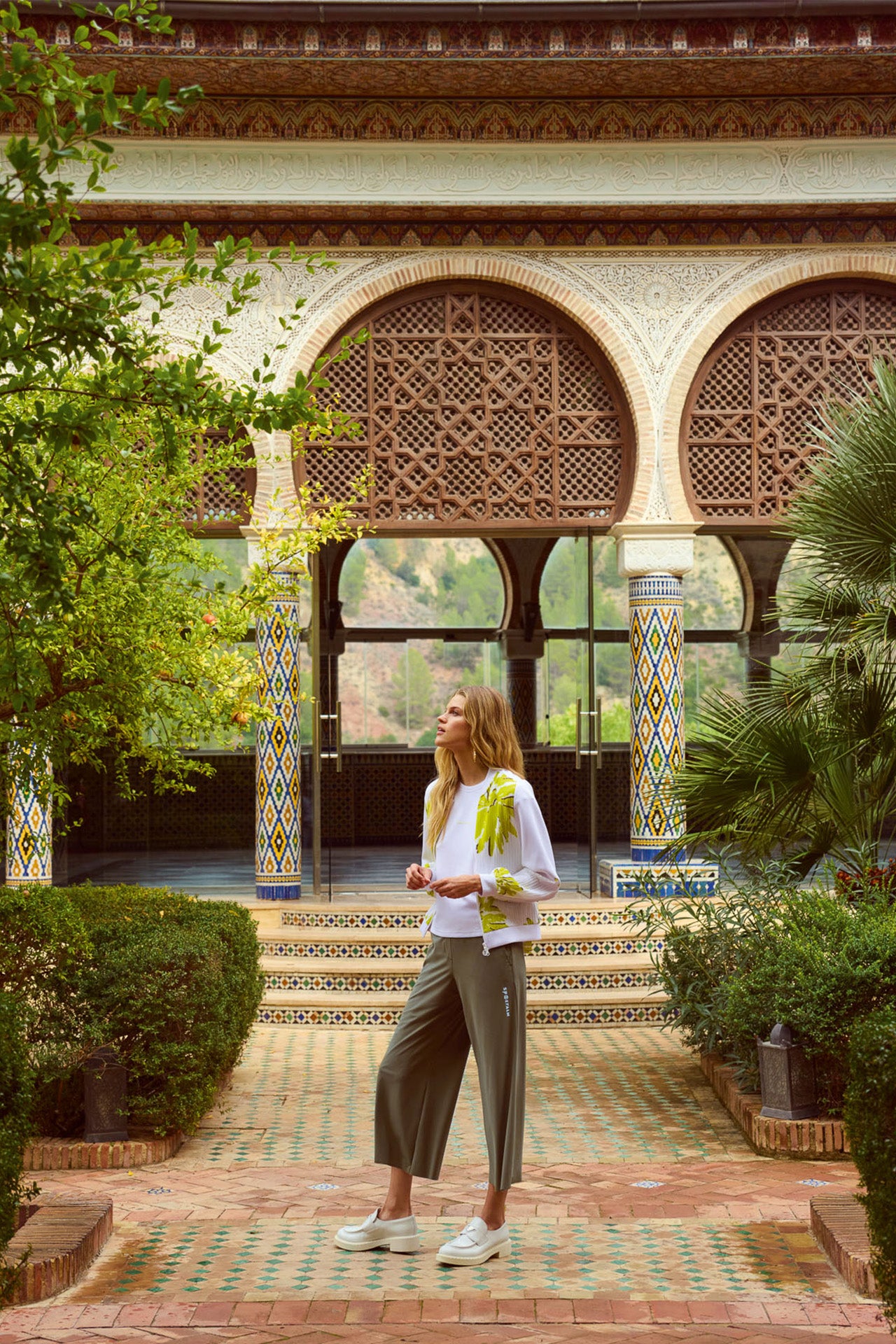 Eine Frau in einem weißen Oberteil und grauen Hosen steht in einem malerischen, begrünten Innenhof mit orientalischer Architektur.