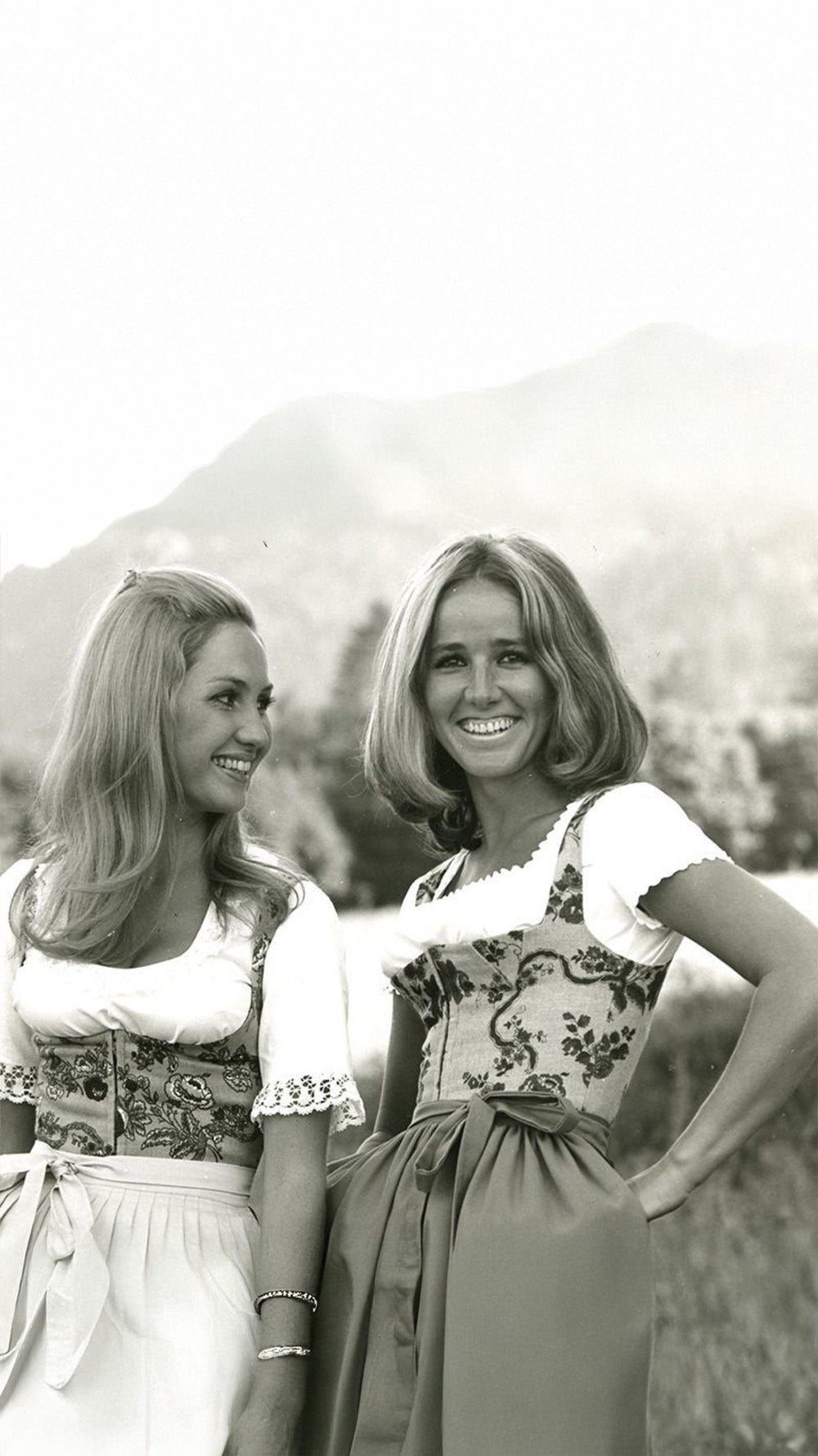 Zwei Frauen in traditioneller bayerischer Tracht stehen vor einer bergigen Landschaft.