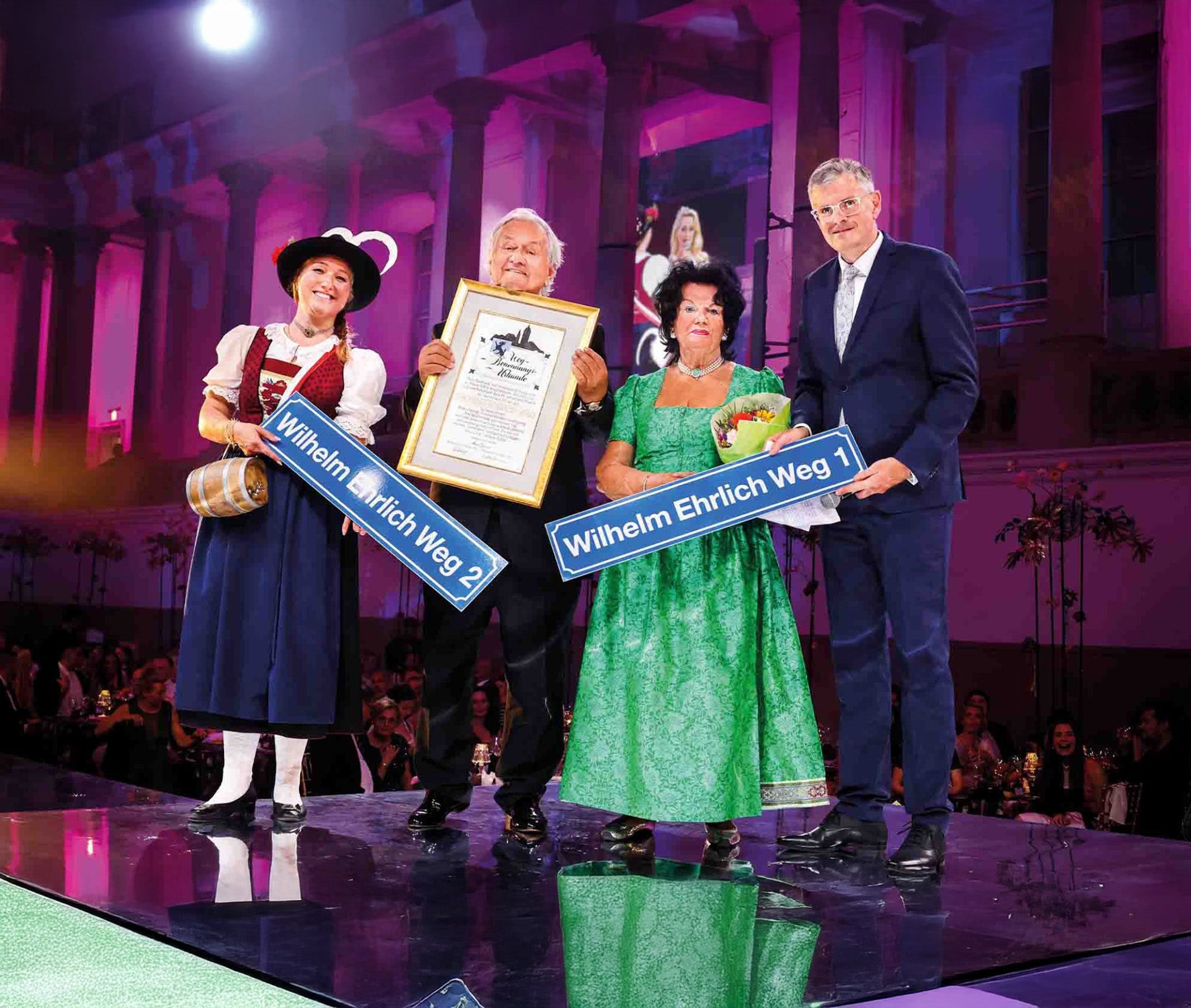 Ein Mann und eine Frau in traditioneller Tracht sowie ein weiteres Paar halten Schilder mit den Aufschriften "Wilhelm Ehrlich Weg 1" und "Wilhelm Ehrlich Weg 3" in einer festlichen Veranstaltung.